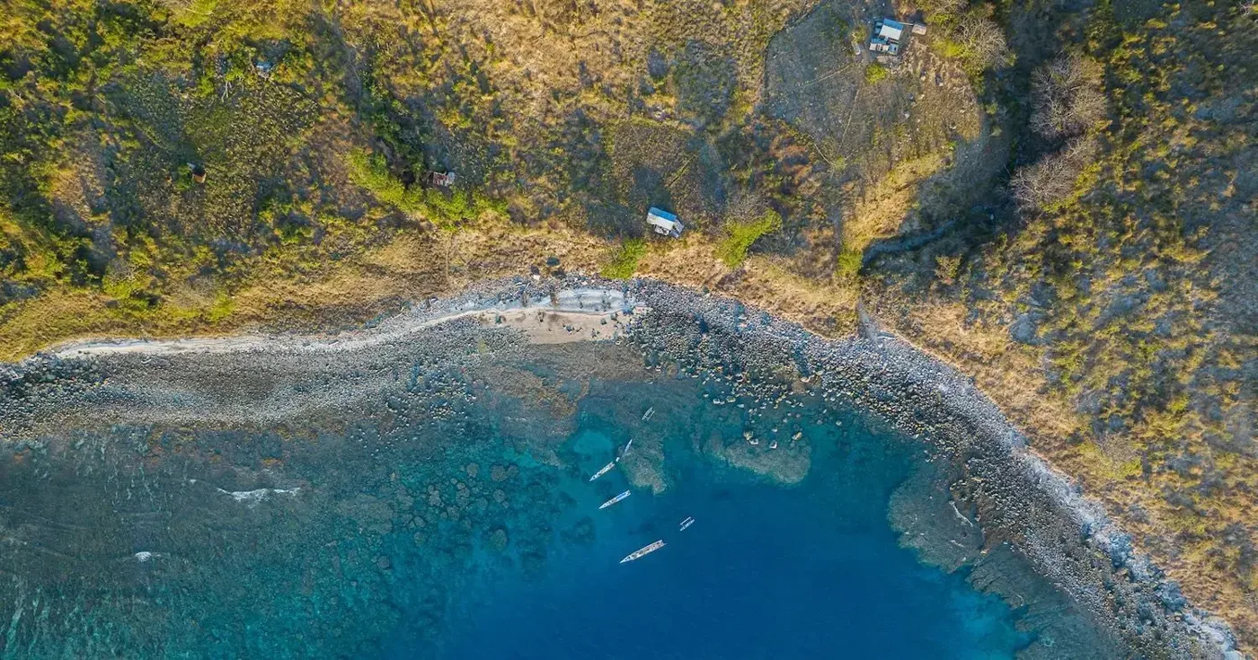 Aerial view of small boats beside a rocky shoreline, clear blue water and scattered buildings among trees