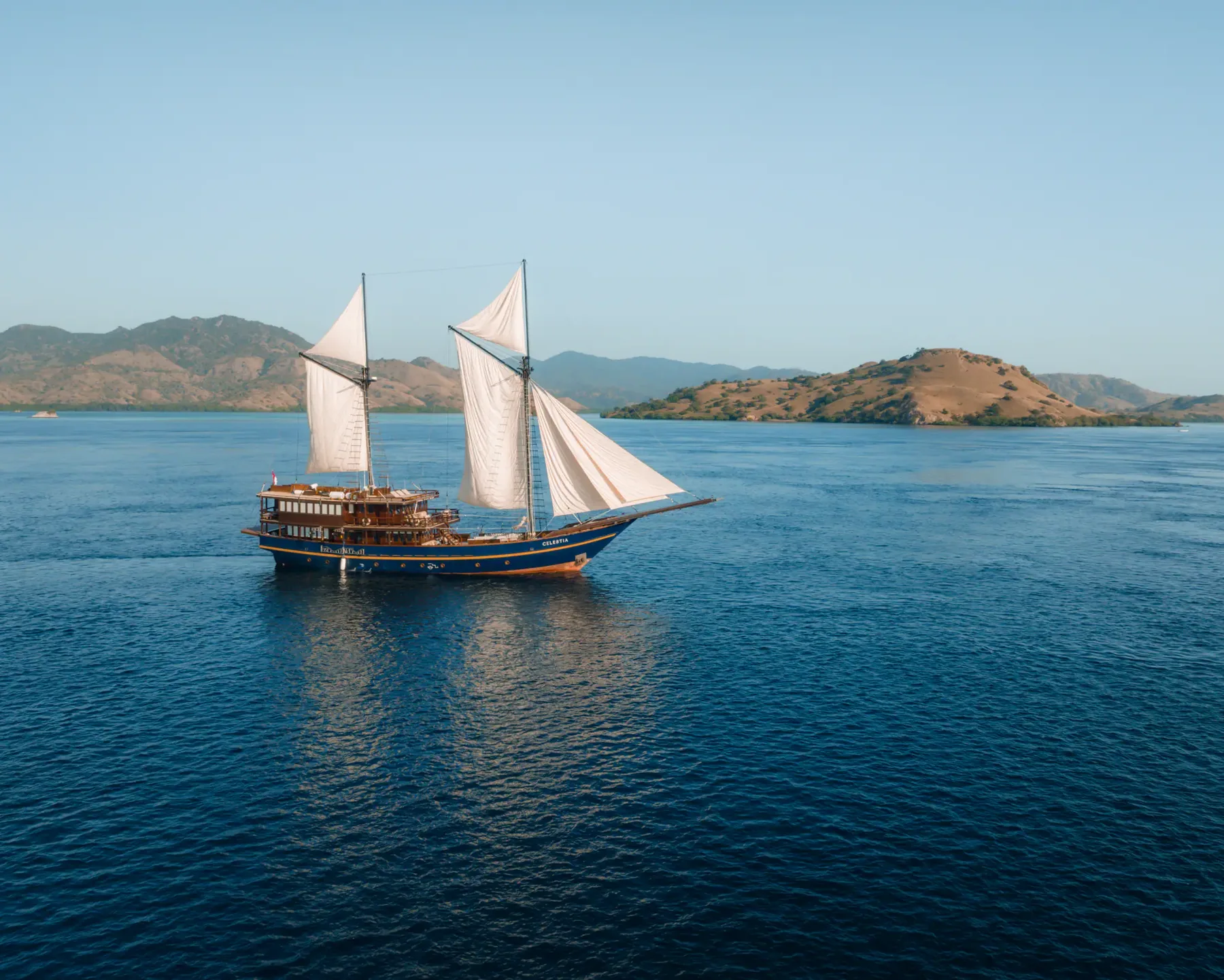 Celestia with white sails raised on blue water beside a hilly island coastline