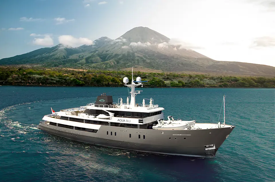 Aqua Blu cruising beside a green island coastline with a volcanic mountain behind it