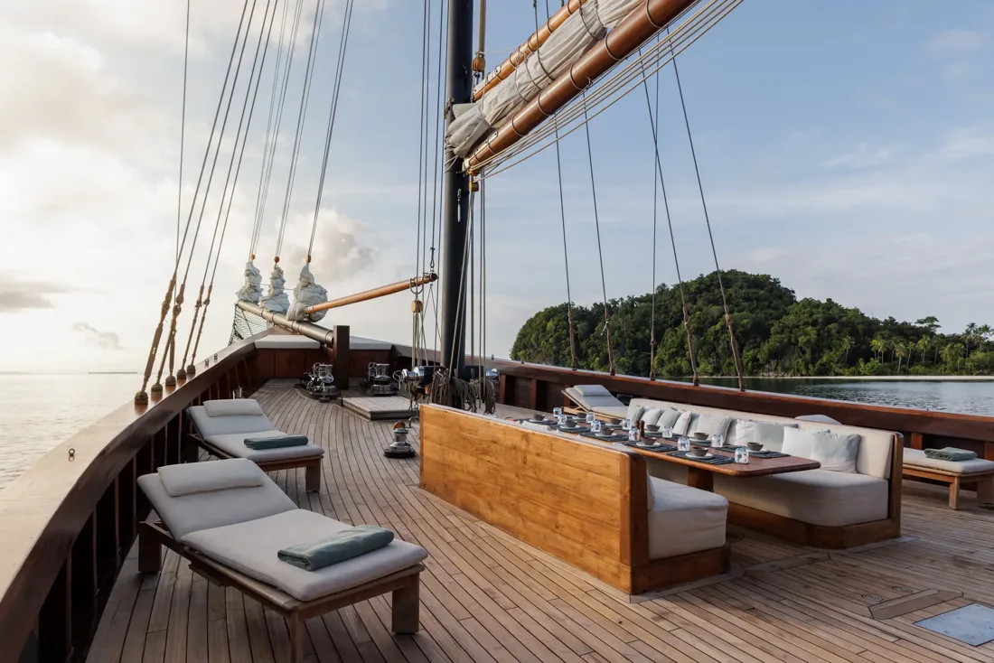 Outdoor dining table and cushioned seating on Vela’s wooden foredeck beside a green island